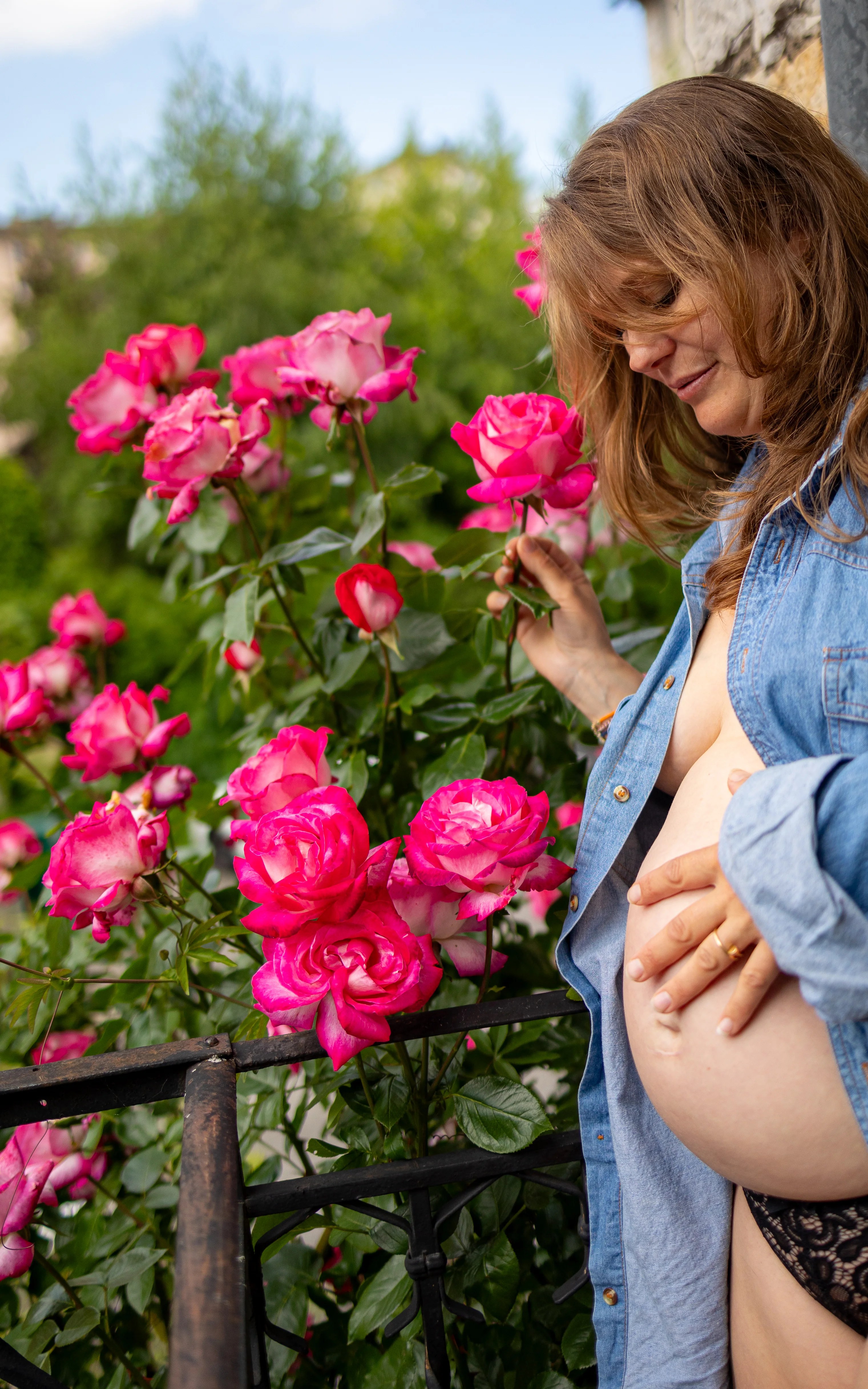 Femme enceinte avec une chemise en jean entrouverte devant un rosier sur un balcon photographiée par Marine Bourely Photographe