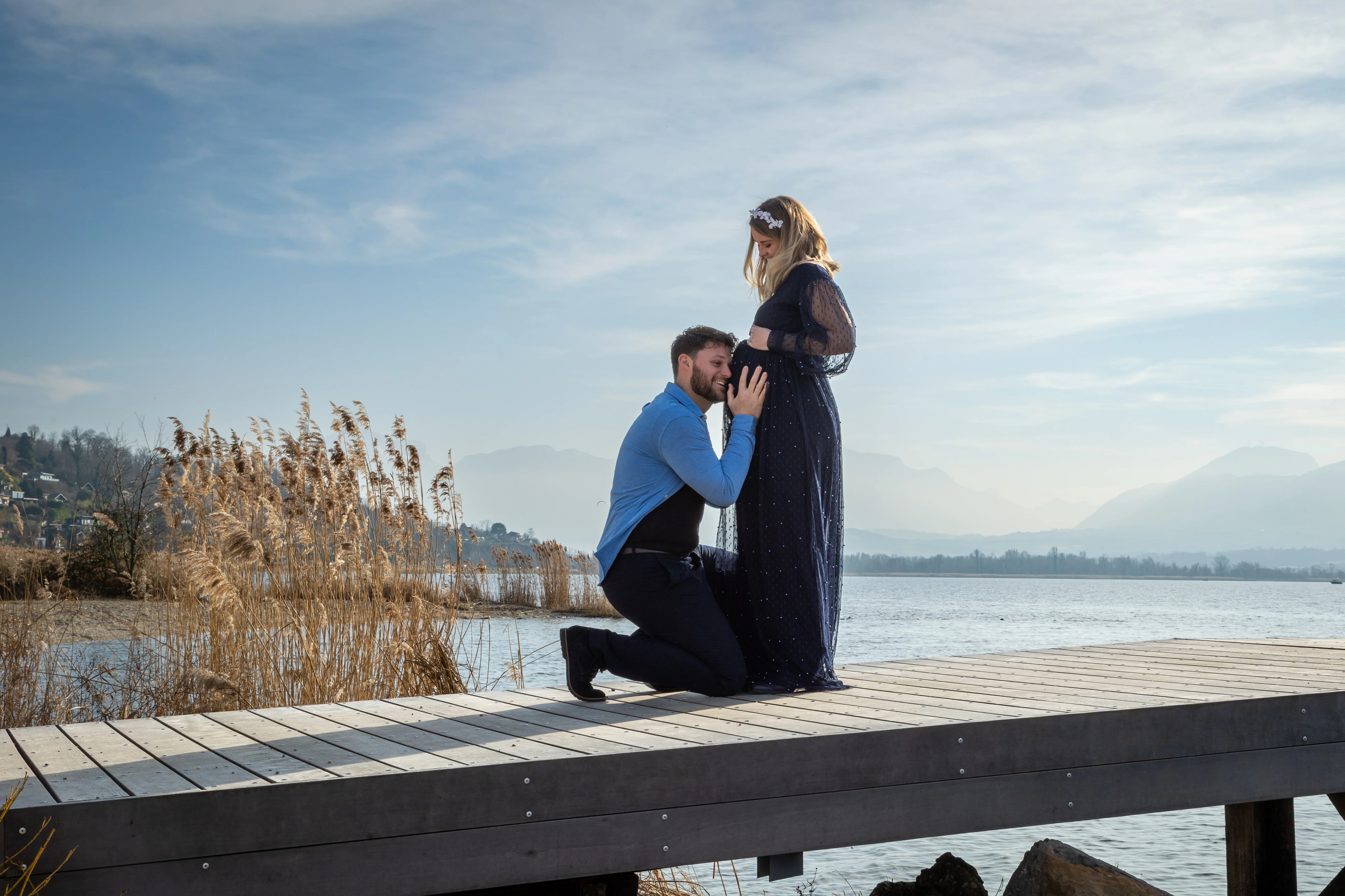 Homme à genou sur un ponton à Aix-les-Bains écoute le ventre de sa femme enceinte photographiés par Marine Bourely Photographe